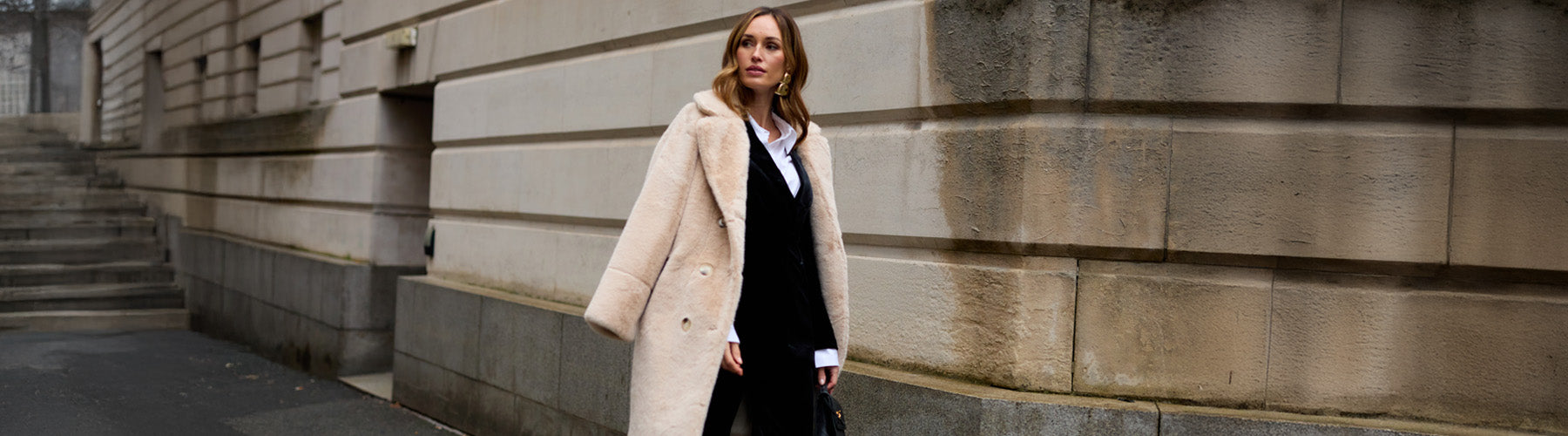 Image showing model wearing the Sienna Tailored Velvet Blazer In black with Sienna Straight Leg Velvet Trouser In Black. The Emma Longline Faux Fur Coat In cream is draped over her shoulder.